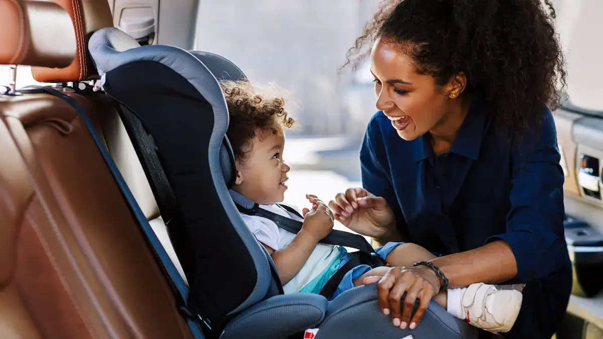A mother putting her child in their safety seat in a car, smiling at her child.