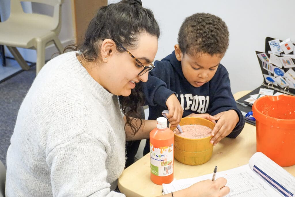 A young child engages in a one-to-one ABA therapy with a therapist, at First Bridge Education Summer School, using colourful educational toys and visual aids to support communication and learning.