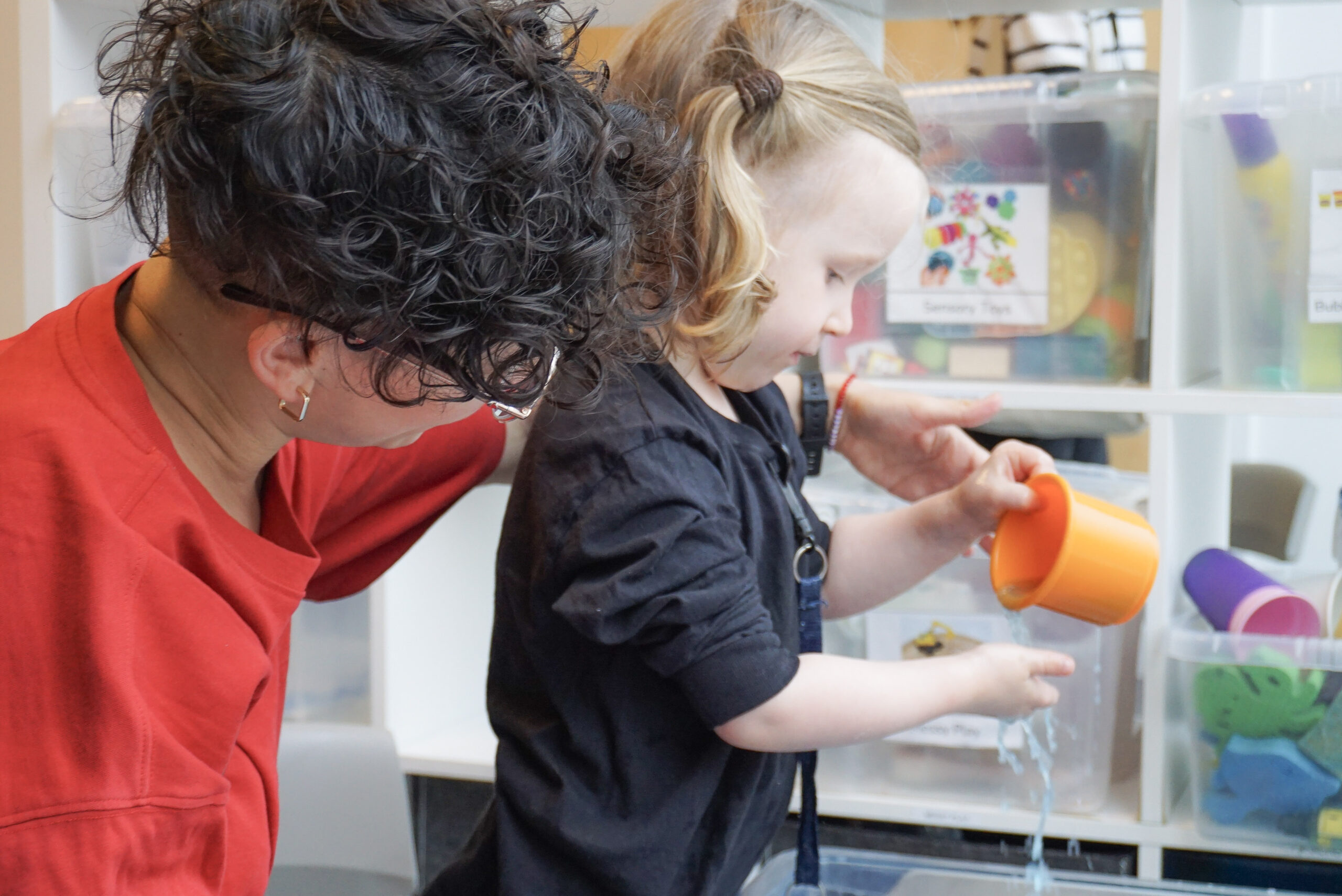 A young autistic child explores water play with an orange cup, supported by an adult at their side. They are engaged in a sensory-rich activity at First Bridge Education, surrounded by labelled storage boxes filled with toys. The environment is calm, structured, and child-centred—supporting the principles of routine, sensory regulation, and connection highlighted in the blog post.