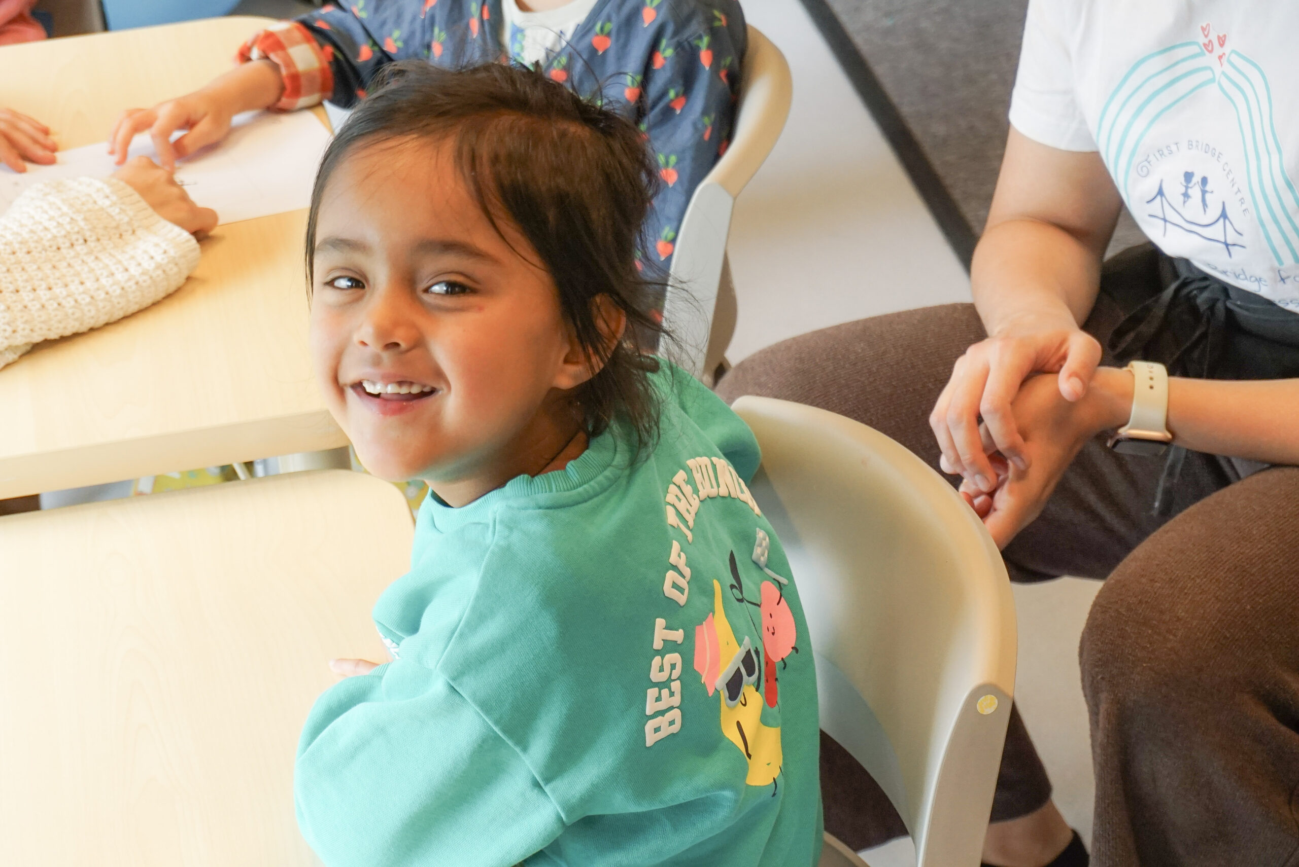 A young child sits at a table in a nursery classroom, smiling joyfully at the camera. Other children and a teacher are nearby, creating a warm and supportive learning environment.