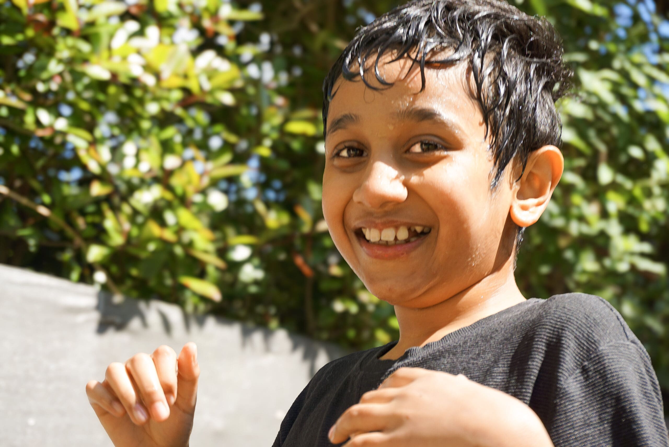 A young child sits on a padded mat while an adult supports her in using a blue AAC (Augmentative and Alternative Communication) device. The adult is guiding her hand or showing her how to use the device. Behind them is a bookshelf with children’s books and soft play cushions, indicating a supportive early years or therapy environment.