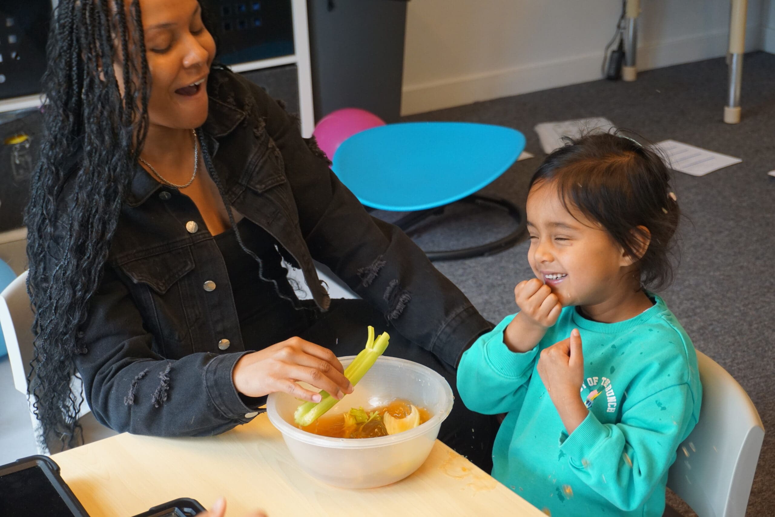 A young boy in nursery sits at a small table with an adult woman, engaging in a playful educational activity using toy animals, including a cow, elephant, and pig. The boy is smiling and appears relaxed and engaged, while a nursery staff member interacts with him supportively in a nurturing early years learning environment.