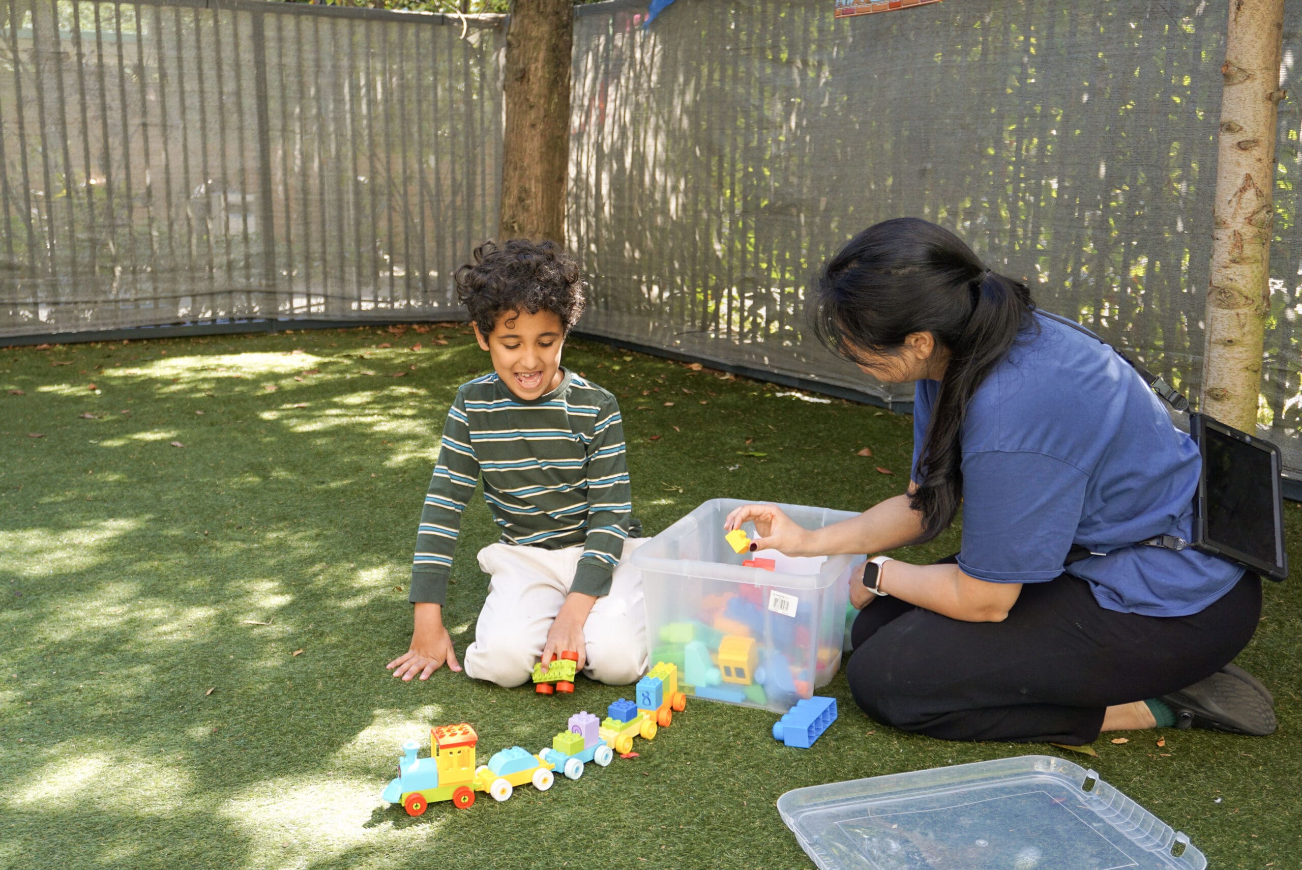 A young boy in nursery sits at a small table with an adult woman, engaging in a playful educational activity using toy animals, including a cow, elephant, and pig. The boy is smiling and appears relaxed and engaged, while a nursery staff member interacts with him supportively in a nurturing early years learning environment.